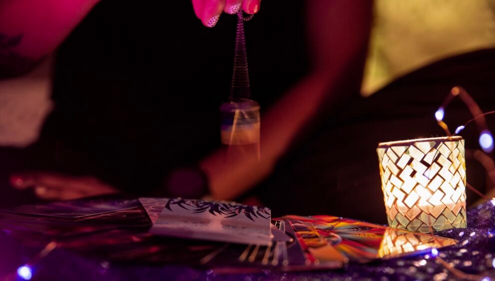 Main balançant un pendule au-dessus de cartes de tarot, avec une bougie allumée à côté.